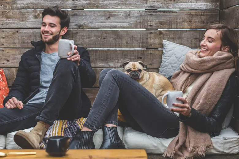 La imagen muestra a una pareja sonriente tomando una bebida caliente, un perro de la raza bulldog está junto a la mujer